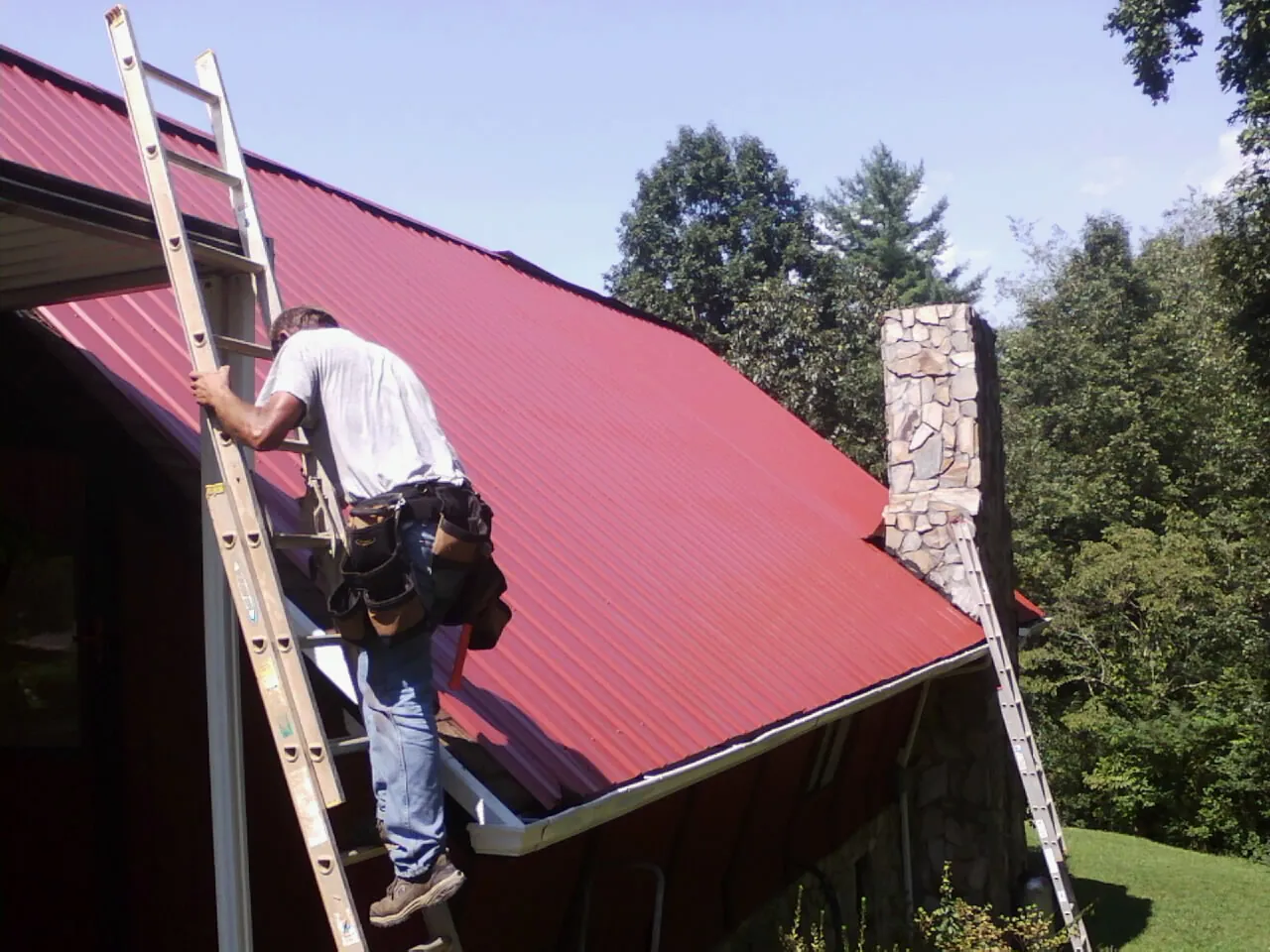 Roofer on a ladder beside a red metal roof and stone chimney — Soffit & Fascia Repair by Roofing Brothers in Upper Sandusky